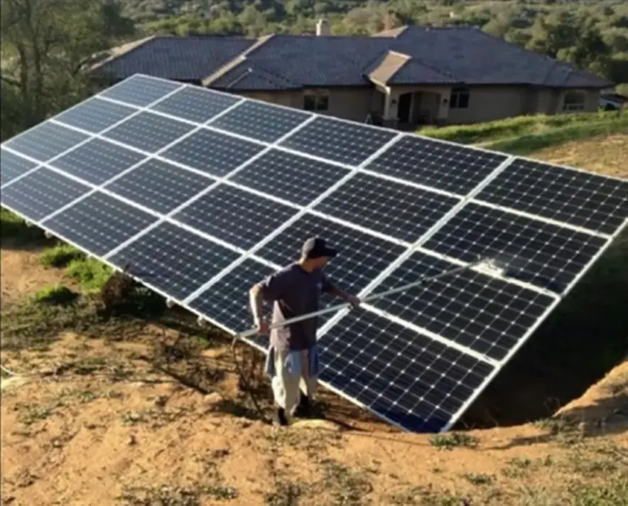 Worker cleaning a large ground-mounted solar panel array with an extension pole