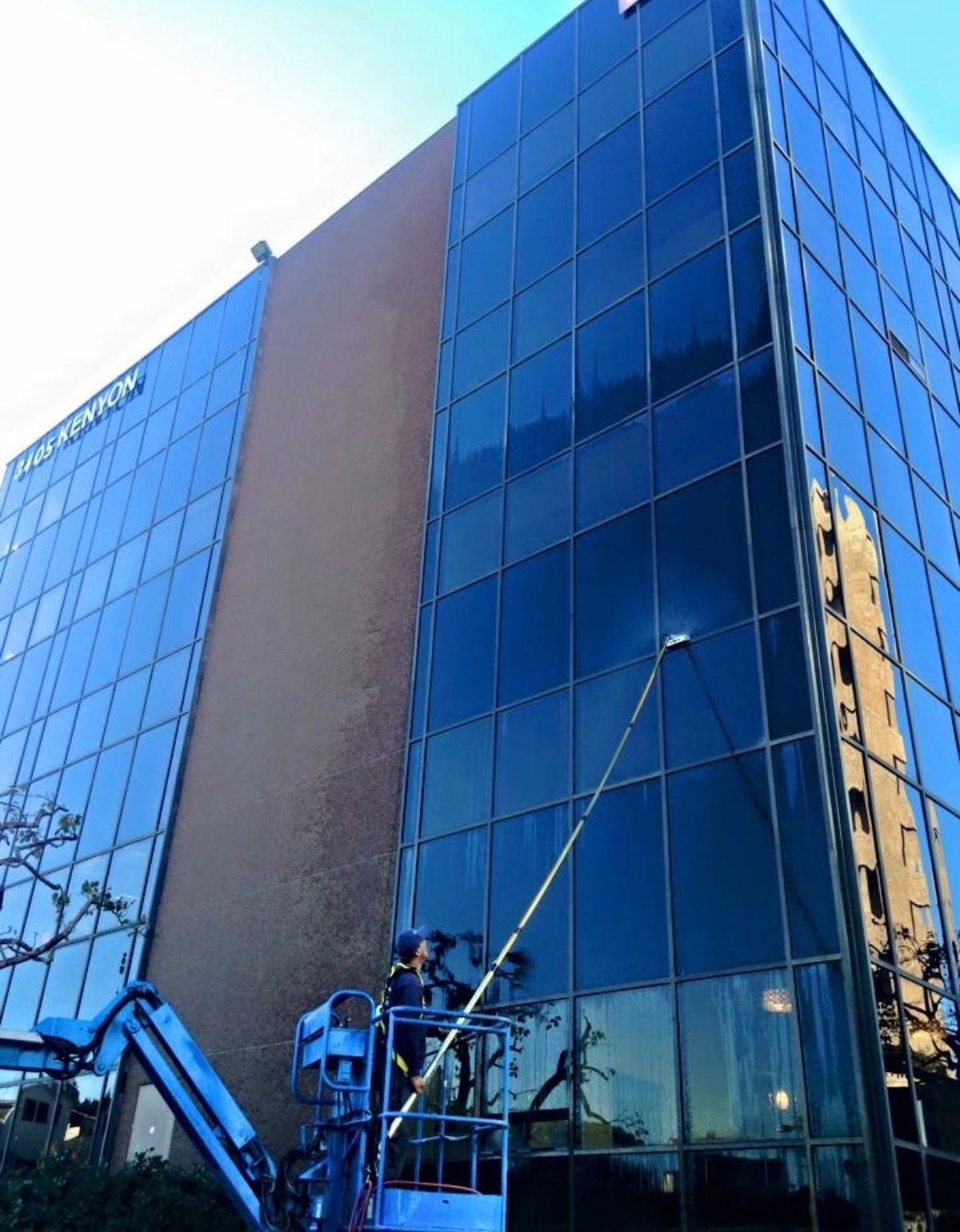 Window cleaner using a long cleaning pole from a blue lift beside a glass-fronted building